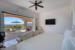 Een slaapkamer met een groot bed, een plafondventilator, een aan de muur bevestigde televisie, en een open terras met lounge fauteuils en parasols.