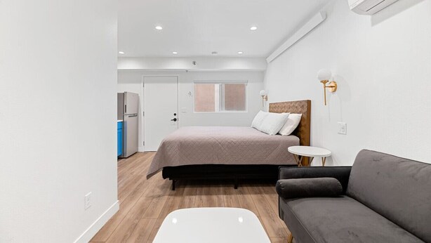 Un dormitorio moderno con cama, sofá gris, una pequeña mesa redonda y una kitchenette al fondo.