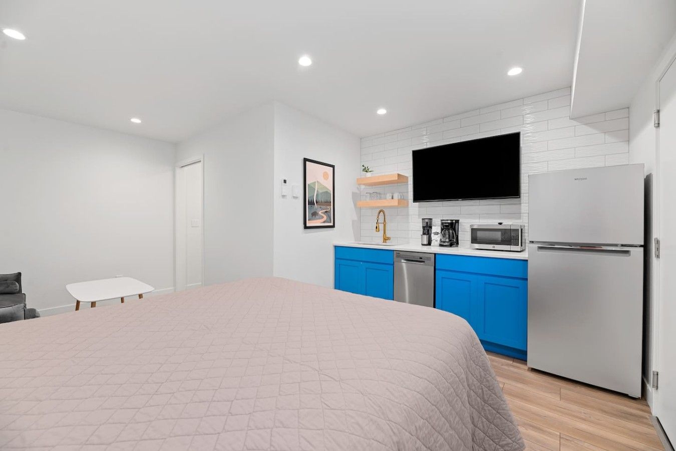 A modern bedroom with a large bed, a small table, and a kitchenette with a refrigerator, microwave, and coffee maker.