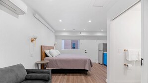 Un dormitorio compacto con cama, una mesita, sofá, ventana y kitchenette.