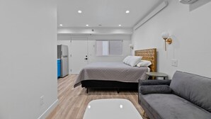 A modern hotel room with a bed, a gray sofa, a small table, a white coffee table, and a kitchenette. - Queen City Inn and Extended Stay #6 (Spearfish)