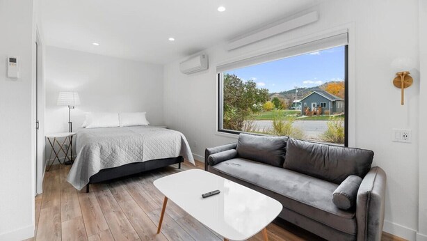 A modern bedroom with a bed, a gray sofa, a small table, and a large window with a view of a house and trees. - Queen City Inn and Extended Stay #1 (Spearfish)