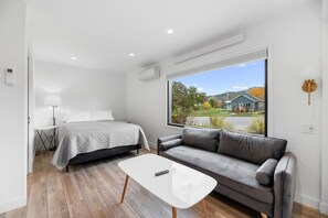 A modern bedroom with a bed, a gray sofa, a small table, and a large window with a view of a house and trees. - Queen City Inn and Extended Stay #1 (Spearfish)