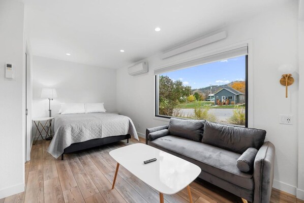 A modern bedroom with a bed, a gray sofa, a small table, and a large window with a view of a house and trees.
