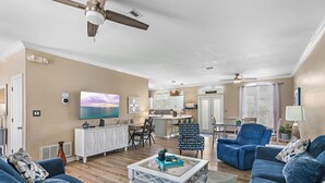 Ein Wohnzimmer mit Sofa, Sessel und Couchtisch. Im Hintergrund befindet sich ein Fernseher, ein Schrank und eine Küchenzeile.