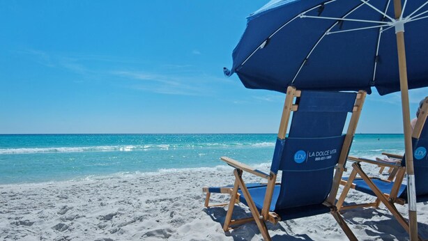 Ein Strandbild mit einem blauen Sonnenschirm und einem Liegestuhl mit der Aufschrift 'La Dolce Vita' am sandigen Ufer.