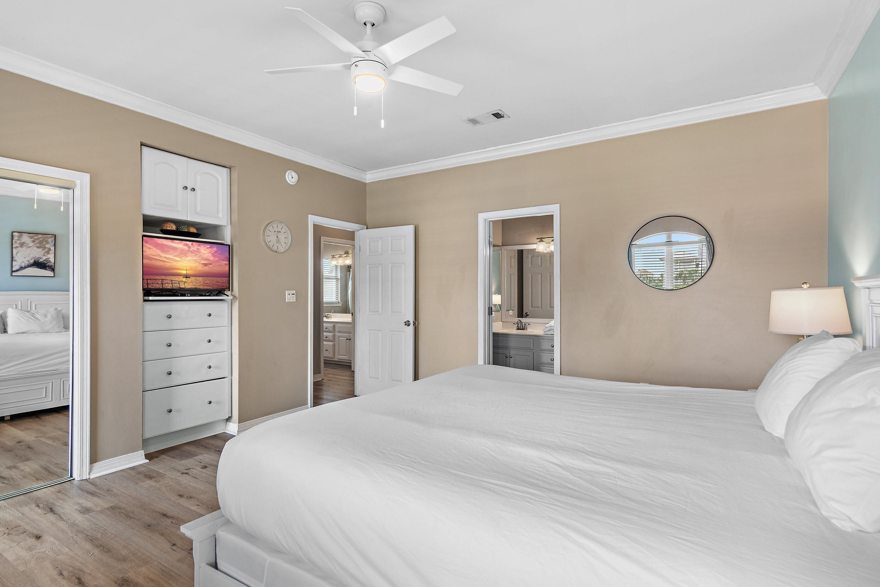 A bedroom with a large bed, a ceiling fan, a TV, and a bathroom visible through an open door.