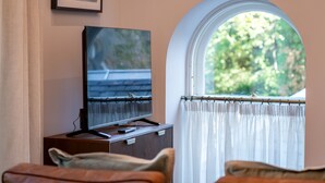 A room with a television on a wooden stand, a window with a view of greenery, and a curtain. - Skye Sands | St Mary’s Gardens (No 7) (St Andrews)
