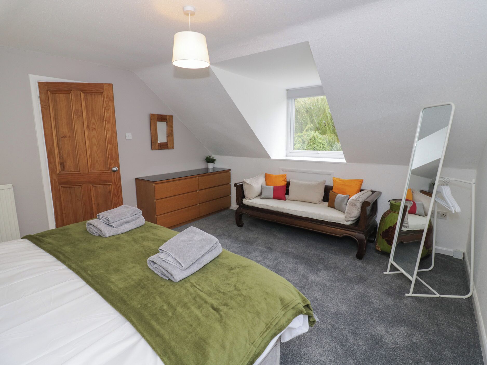 A bedroom with a bed, a dresser, a bench with cushions, a mirror, and a skylight.
