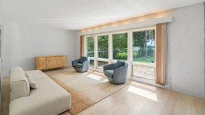 A spacious living room with a large window, a sofa, two armchairs, and a wooden cabinet.