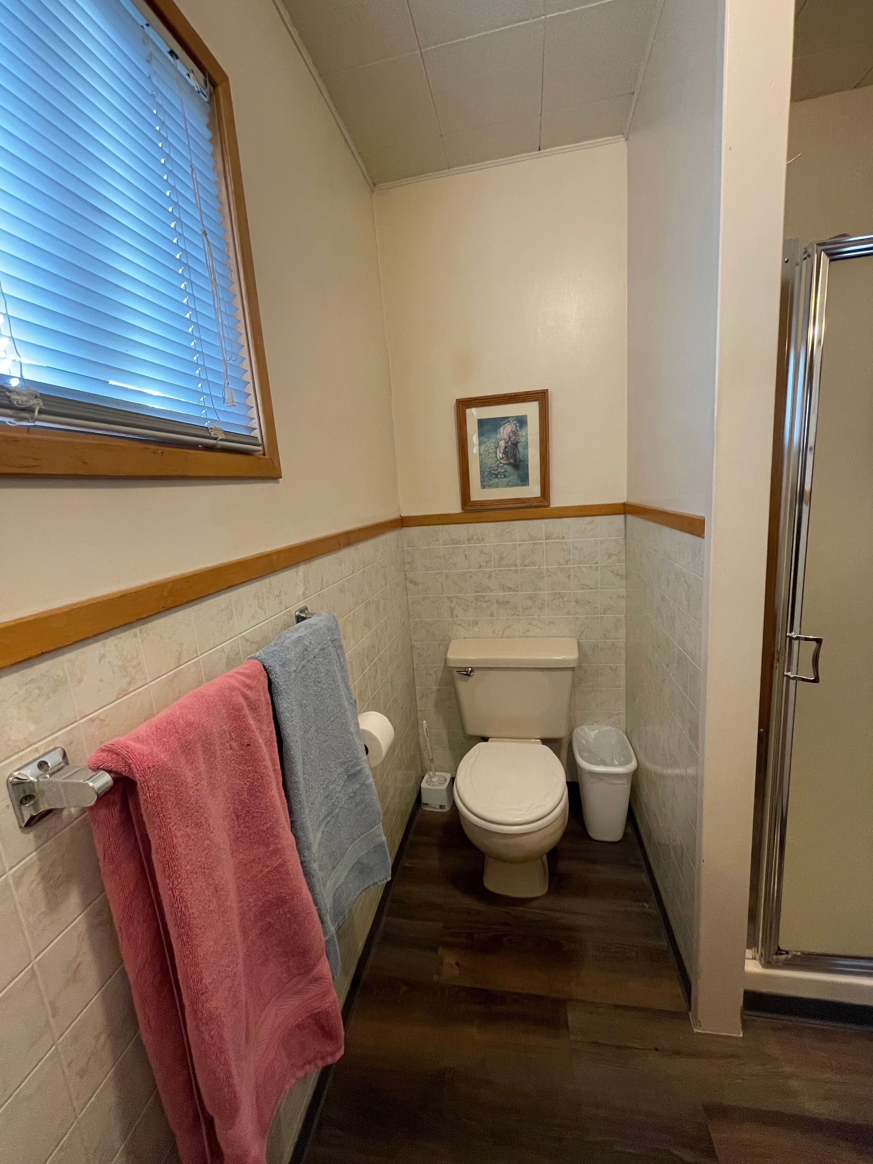 Une petite salle de bain avec des toilettes, une poubelle et une fenêtre avec des stores.