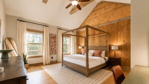 Een slaapkamer met een hemelbed, houten balken in het plafond, een groot raam met gordijnen, een bureau en een stoel.