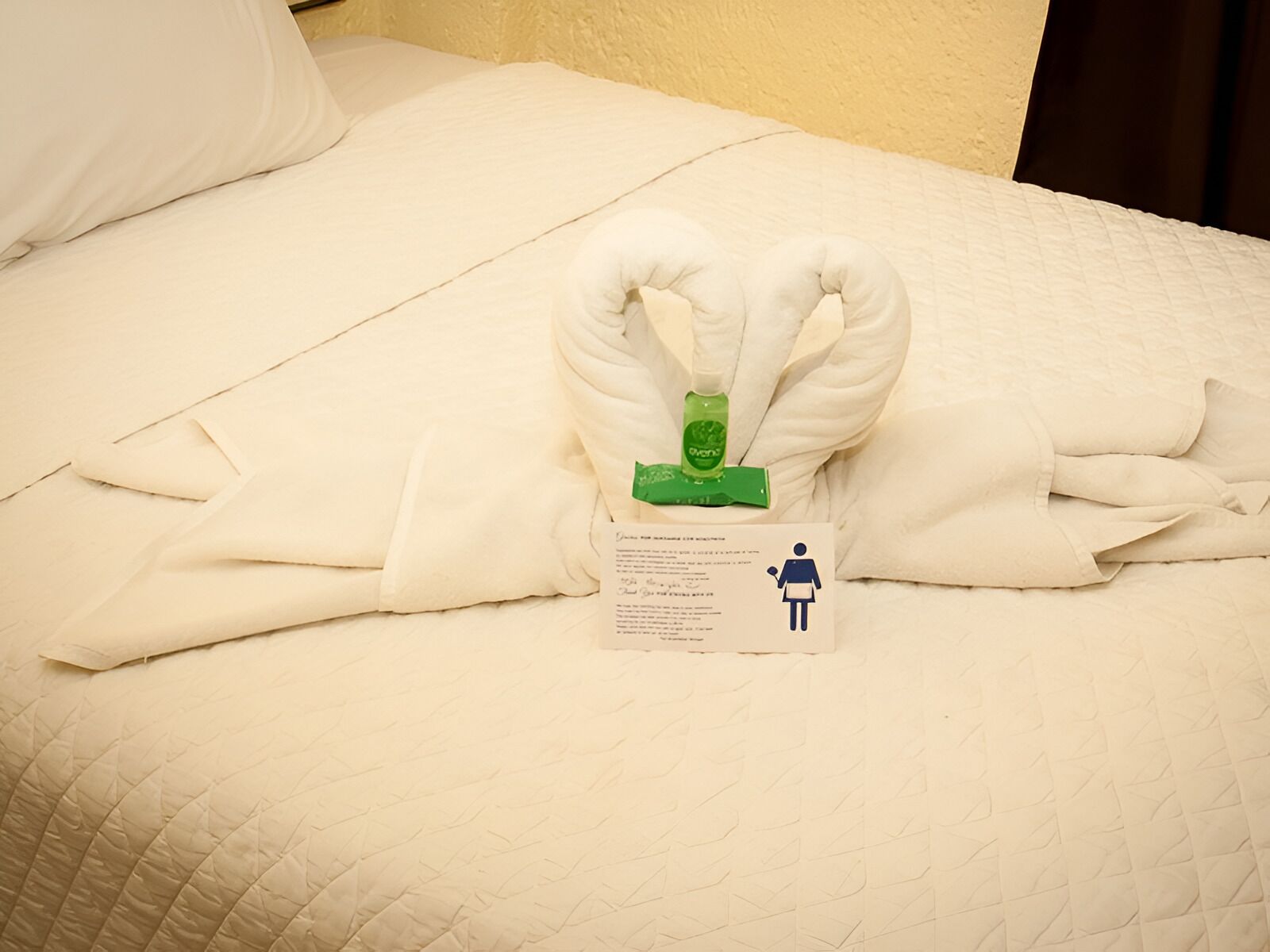 Un letto rifatto con cura, un asciugamano piegato e un cartoncino con il simbolo del bagno.