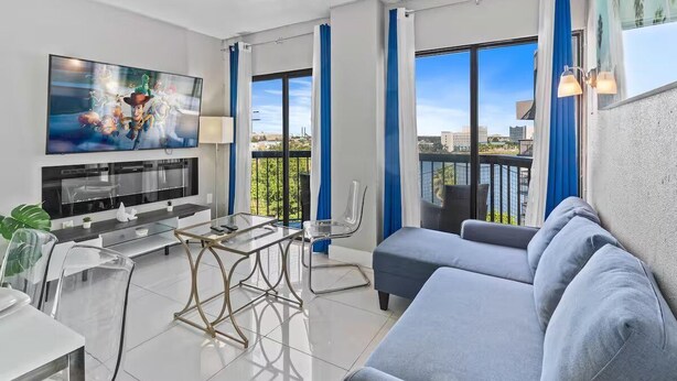 A modern living room with a flat-screen TV, a sofa, a glass coffee table, and a dining area with chairs. - Modern 2-Bedroom Enclave Condo Retreat (Orlando)