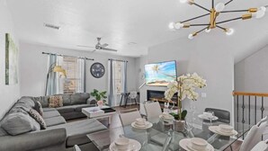 A modern living room with a glass dining table set for four, a gray sofa, a ceiling fan, and a flat-screen TV displaying a beach scene.