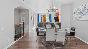 A modern dining area with a glass table and chairs, a chandelier, and a view into the kitchen.