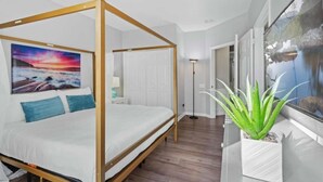 A four-poster bed with a painting of a sunset, a potted plant, and a view of a mountain through a window.