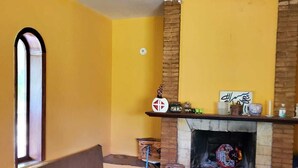 A room with a yellow wall, a fireplace, a bench, and a window. - Charming place, with a cozy single-story house. (Mairinque)