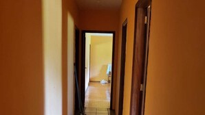 A hallway with orange walls and tiled flooring, leading to a room with a bed and a chair. - Charming place, with a cozy single-story house. (Mairinque)