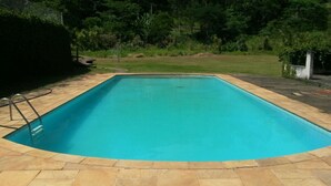 A rectangular swimming pool with a tiled deck, surrounded by a grassy area and trees. - Charming place, with a cozy single-story house. (Mairinque)