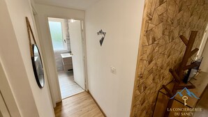 Un pasillo con piso de madera, un espejo a la izquierda, un baño visible a través de una puerta abierta, y una pared con diseño geométrico y un logotipo.