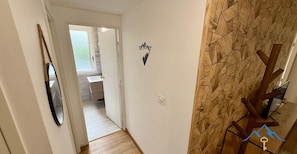 A hallway with a wooden floor, a mirror on the left, a bathroom visible through an open door, and a wall with a geometric pattern and a logo. - Le Bélouga 4 Le Mont Dore (Mont-Dore)