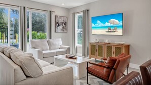 A modern living room with a large TV displaying a beach scene, a white sofa, a coffee table, and a leather armchair. - Inviting Townhome near Disney with Resort & Waterpark Access - 7833SD (Kissimmee)