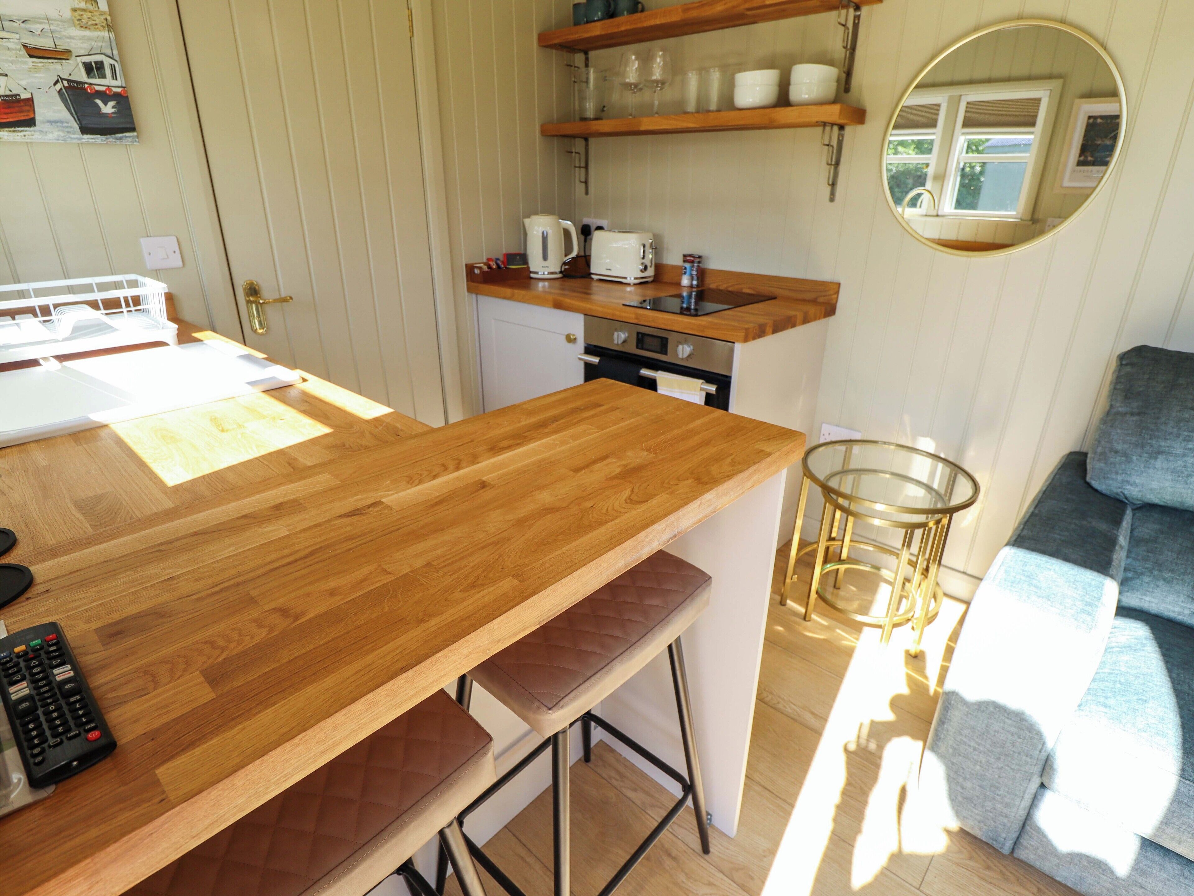 A kitchen with wooden countertops, a coffee maker, and a stove. There is a glass table and a grey sofa in the living area.