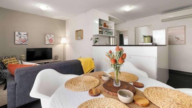 A modern living room with a sofa, a dining table with placemats, a TV, and a kitchen area with cabinets and a microwave.