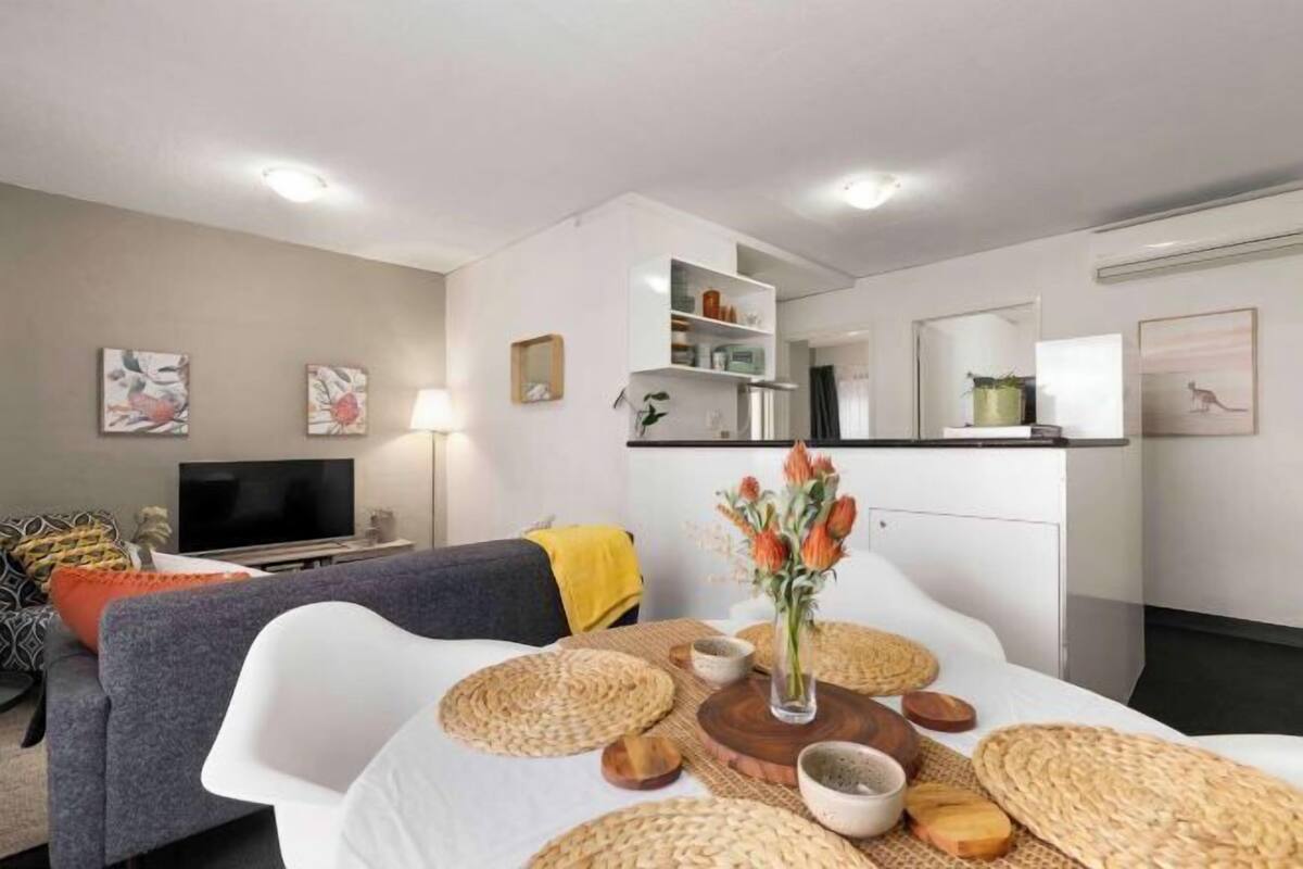 Una sala moderna con un sofá, una mesa de comedor con individuales, un televisor y una zona de cocina con armarios y un microondas.