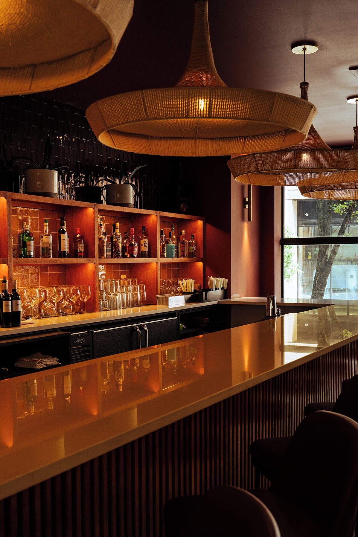 A bar area with shelves of bottles and glasses, a long counter, and hanging lamps.