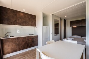 A compact kitchen with a sink, stove, and refrigerator, a dining area with a table and chairs, and a bedroom with a bed and bedside table. - Mobile Home 'Camping Belsito Evo Medium 511' with Shared Pool, Wi-Fi & Air Conditioning (Vico)