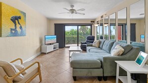 A modern living room with a sofa, a wooden chair, a small table, and a ceiling fan. - Las Brisas 502 (New Smyrna Beach)