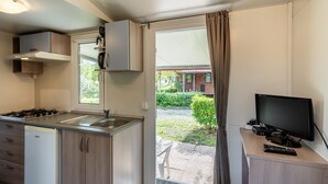 Een compacte keuken met een gasfornuis, spoelbak en een kleine koelkast. Er is een televisie op een houten standaard en een deur naar een buitenruimte met uitzicht op een tuin.