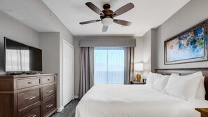 Quarto de hotel com cama, televisão, escrivaninha com gavetas, ventoinha no teto, quadro na parede e janela com cortinas.