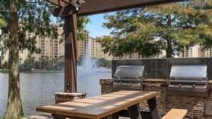 Eine Terrassengaststätte mit Grill und Tisch, mit Blick auf ein Gewässer und einen Gebäudekomplex.