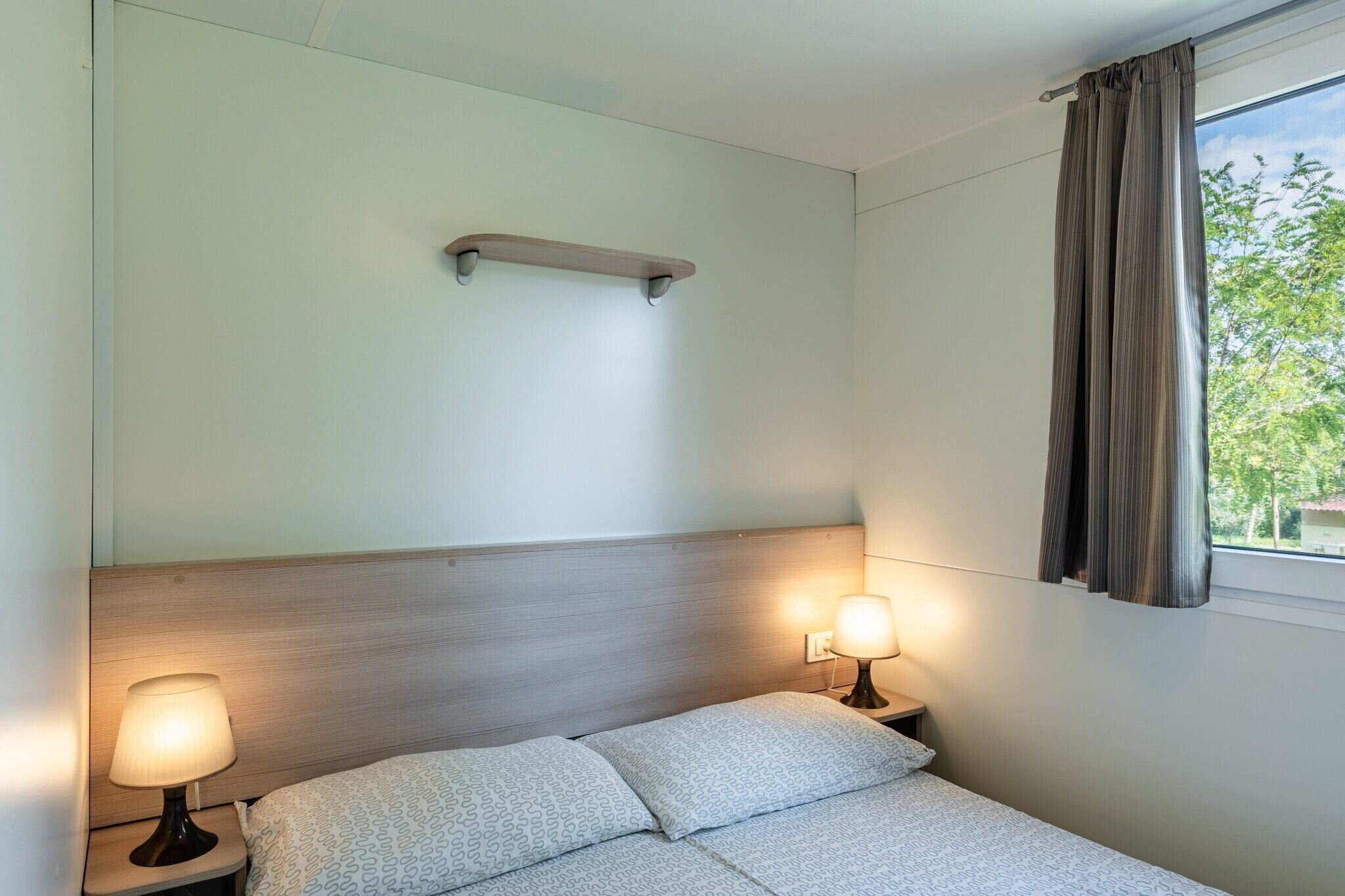 Een hotelkamer met een bed, twee bedlampen, een houten hoofdeinde, een plank boven het bed en een raam met gordijnen.