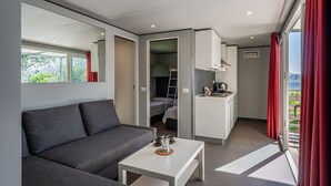 A modern room with a grey sofa, a small white table, a kitchenette, and a view of greenery through large windows.