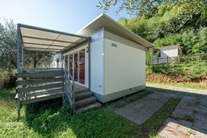 A small, single-story building with a covered entrance, a set of steps leading to a door, and a pathway leading to a small shed in the background.