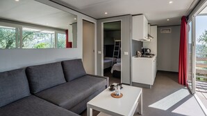 Une chambre moderne avec un canapé gris, une petite table blanche, un coin cuisine et un balcon agrémenté de rideaux rouges.