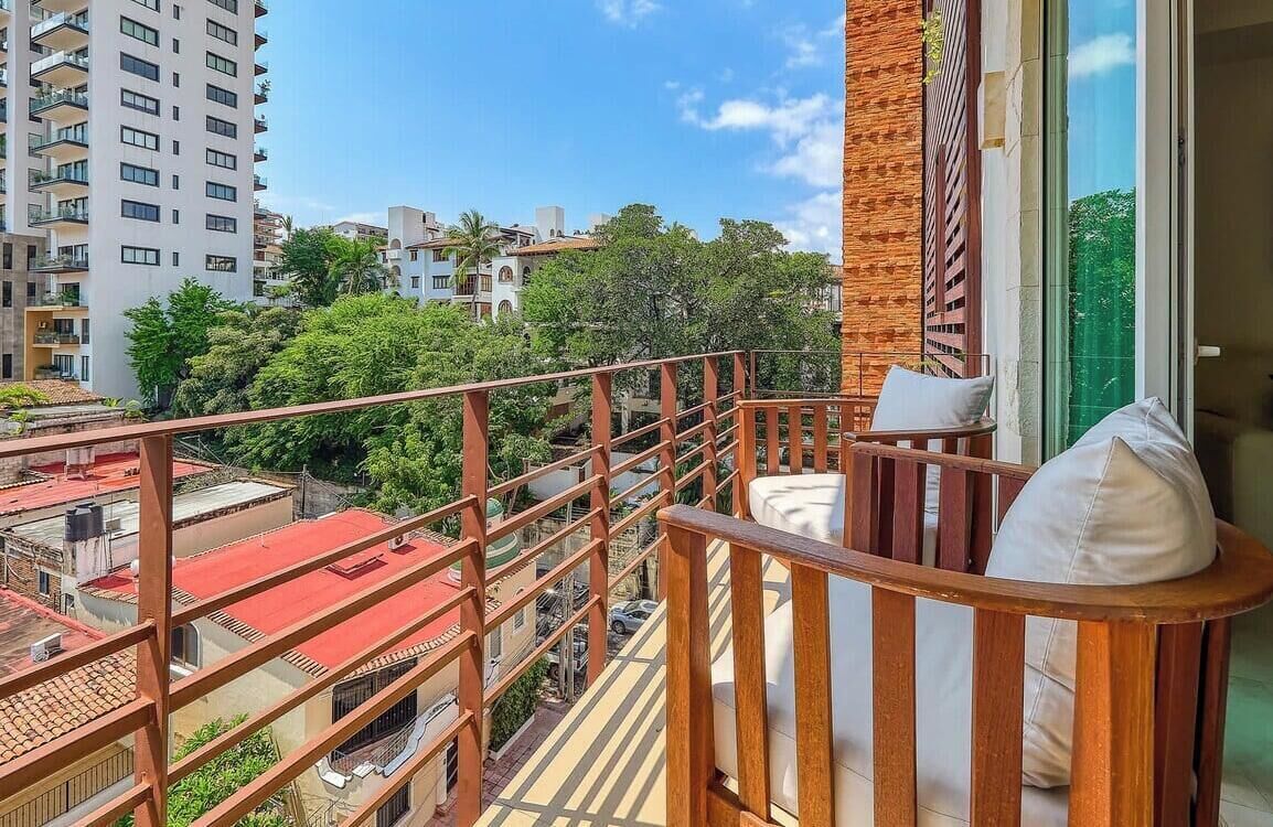A balcony with a wooden railing, a cushioned seat, and a view of surrounding buildings and greenery.