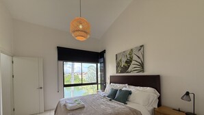 A bedroom with a large bed, a nightstand, a lamp, a window with a view, and a hanging light fixture. - Brand New lake view Penthouse with A/C, pool, gym, tennis in common area (Río Oro)