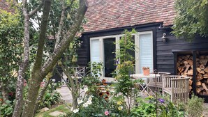 Ett rustikt trähus med ett tak täckt av tegelpannor, en trädgård med olika växter och blommor samt en liten uteplats med ett bord och stolar.