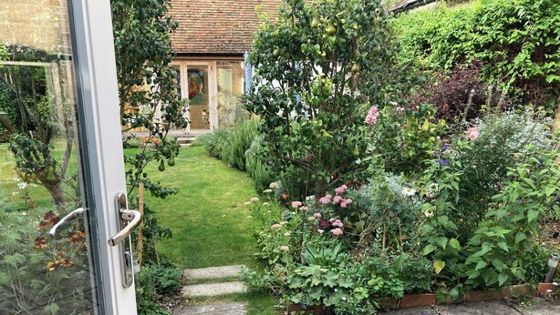 En trädgårdstig som leder fram till ett hus med ett tak av tegel, omgivet av olika växter och blommor.