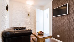A modern living room with a leather sofa, a wooden coffee table with fruit, a stone-textured wall, a white door, and a mounted TV. - High-quality, centrally located apartment in the middle of the city (Rudolstadt)