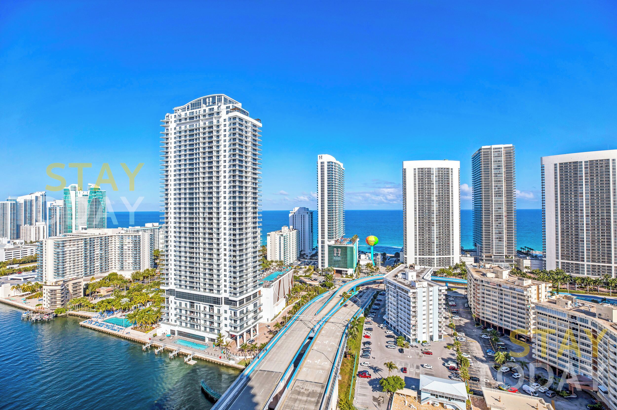 A high-rise hotel with a view of the ocean, a parking area, and a swimming pool.