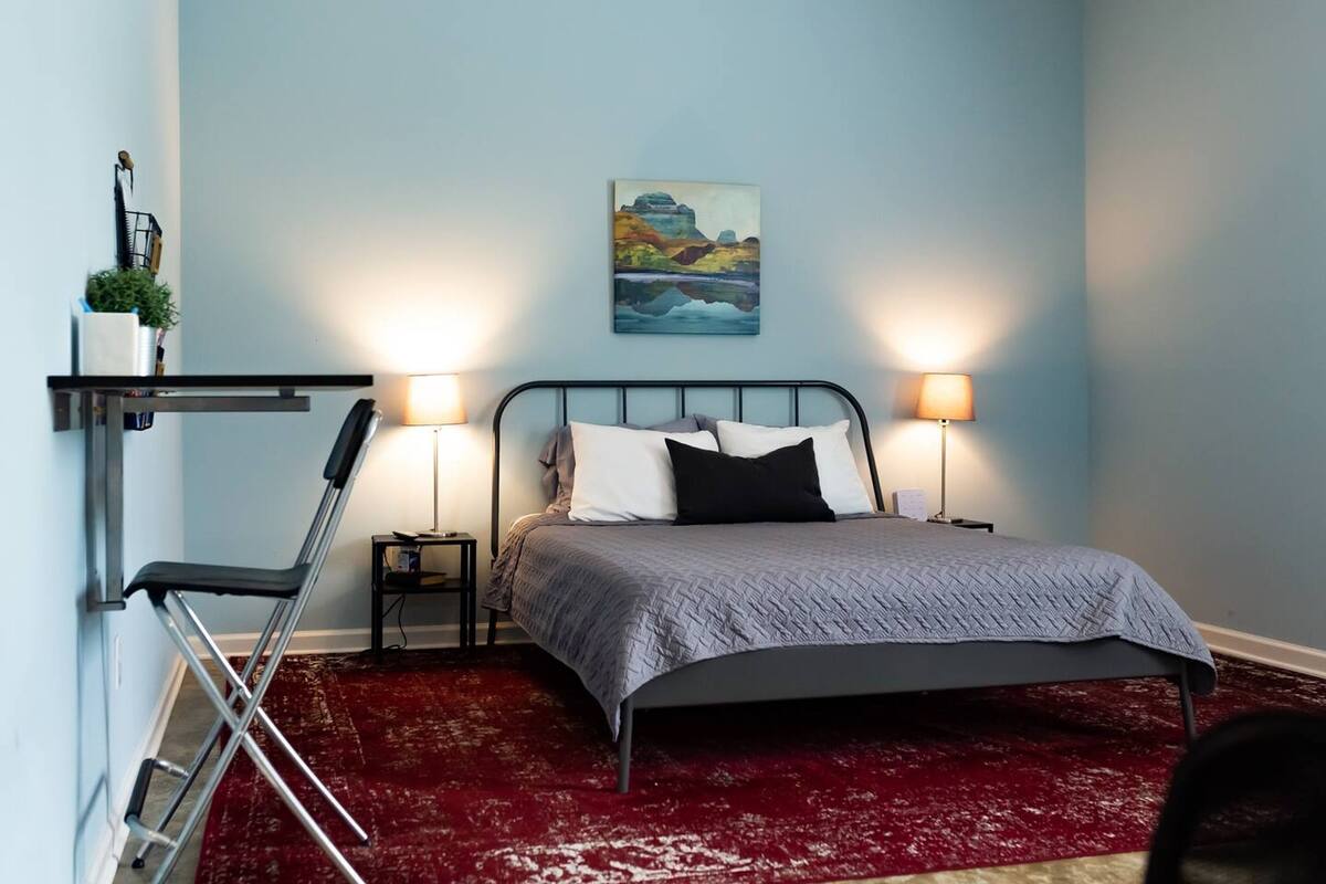 A bedroom with a bed, two bedside lamps, a red carpet, a wall-mounted shelf with a plant, and a painting.
