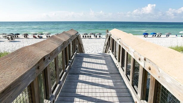 Ein hölzerner Steg führt zu einem Strand mit Sonnenschirmen und Liegestühlen.