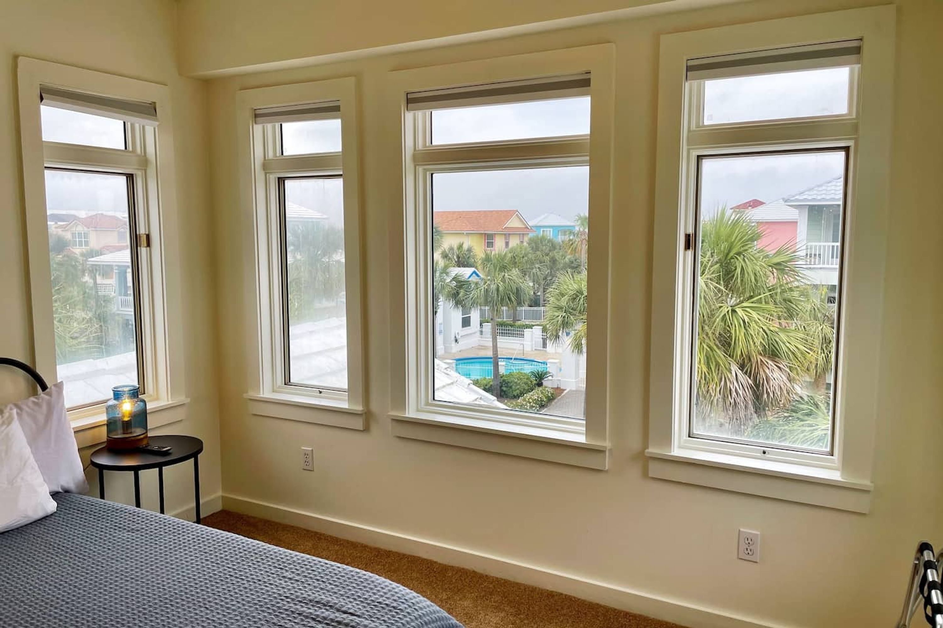 A bedroom with three large windows, a bed, a small table with a lamp, and a view of a swimming pool and buildings outside.