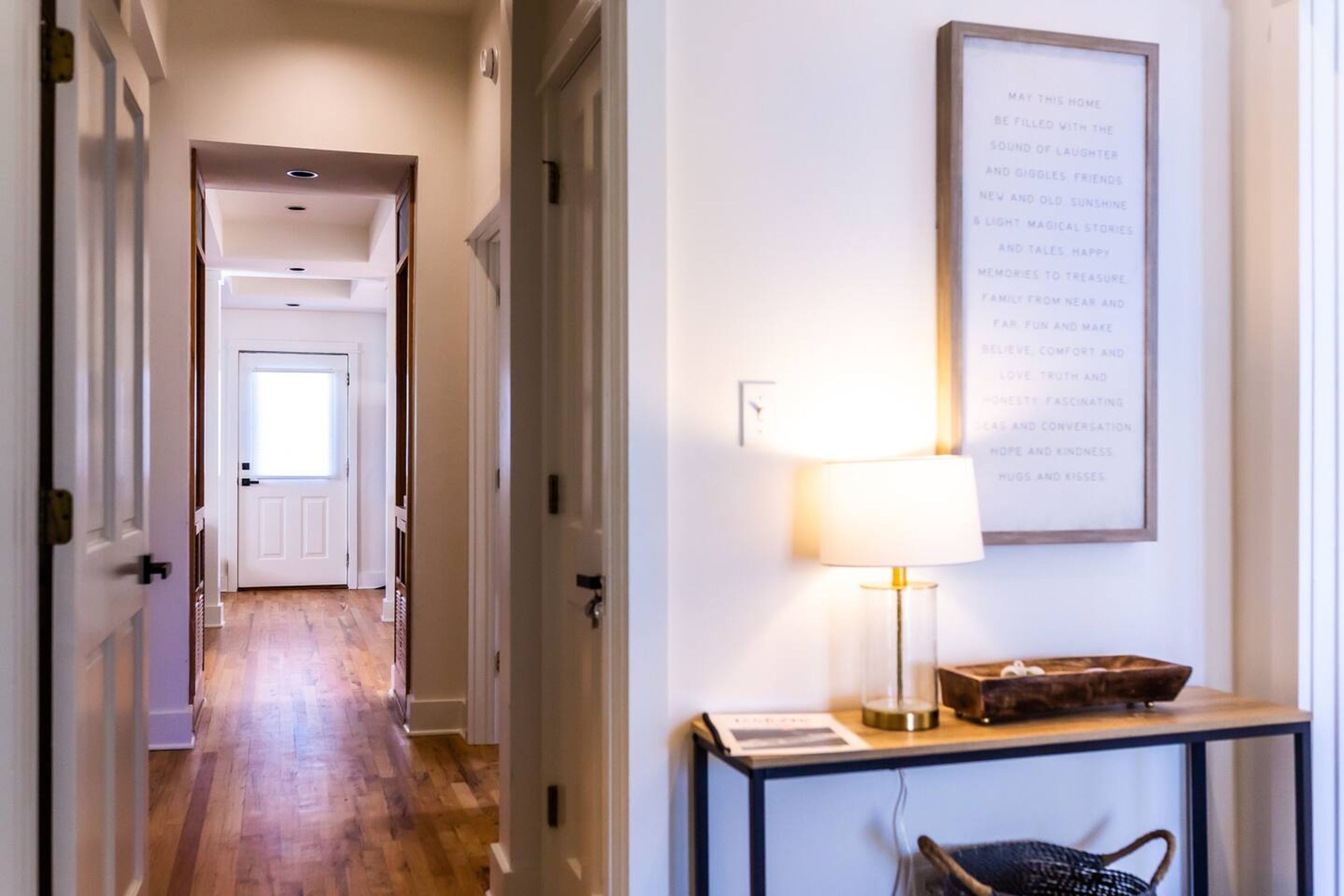 A hallway with wooden flooring and white walls, leading to a door at the end. There is a framed quote on the wall and a table with a lamp and a decorative box.